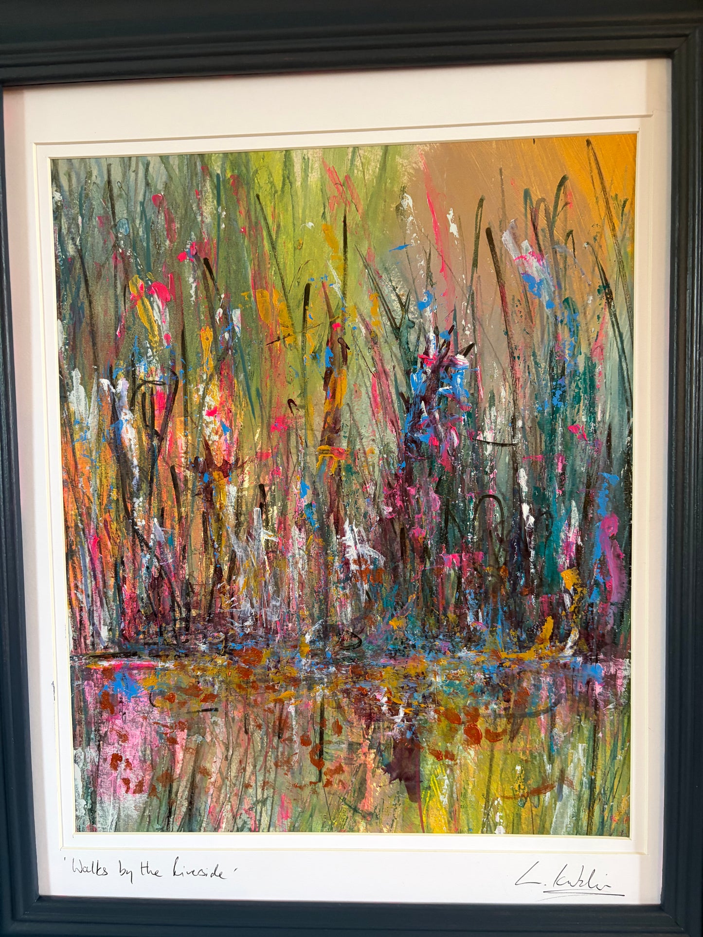 A framed abstract landscape painting depicting tall grasses and reeds with colorful brushstrokes of green, yellow, pink, orange, blue, and brown, presented in a black frame with a white mat.
