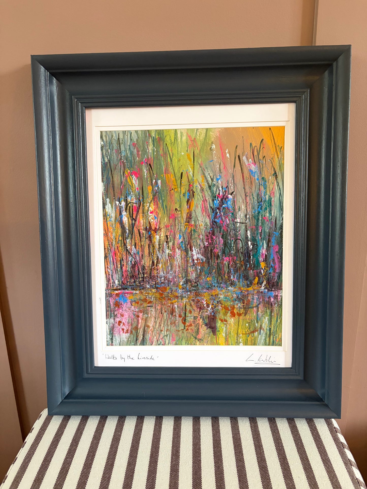 A framed abstract landscape painting featuring colorful brushstrokes of green, yellow, pink, blue, and orange, housed in a dark slate blue frame and displayed against a tan wall.