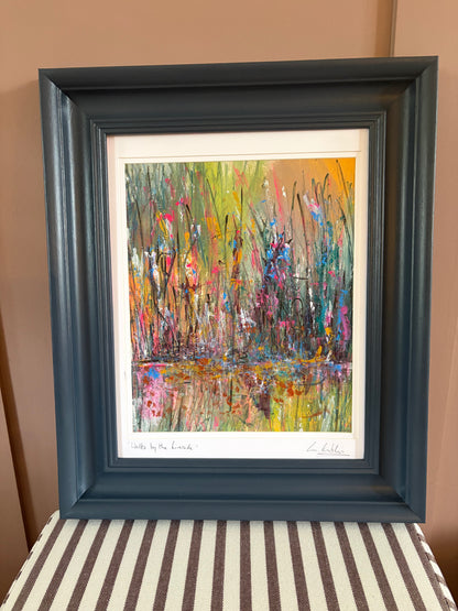A framed abstract landscape painting featuring colorful brushstrokes of green, yellow, pink, blue, and orange, housed in a dark slate blue frame and displayed against a tan wall.