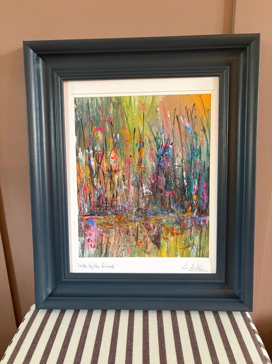 A framed abstract landscape painting featuring colorful brushstrokes of green, yellow, pink, blue, and orange, housed in a dark slate blue frame and displayed against a tan wall.
