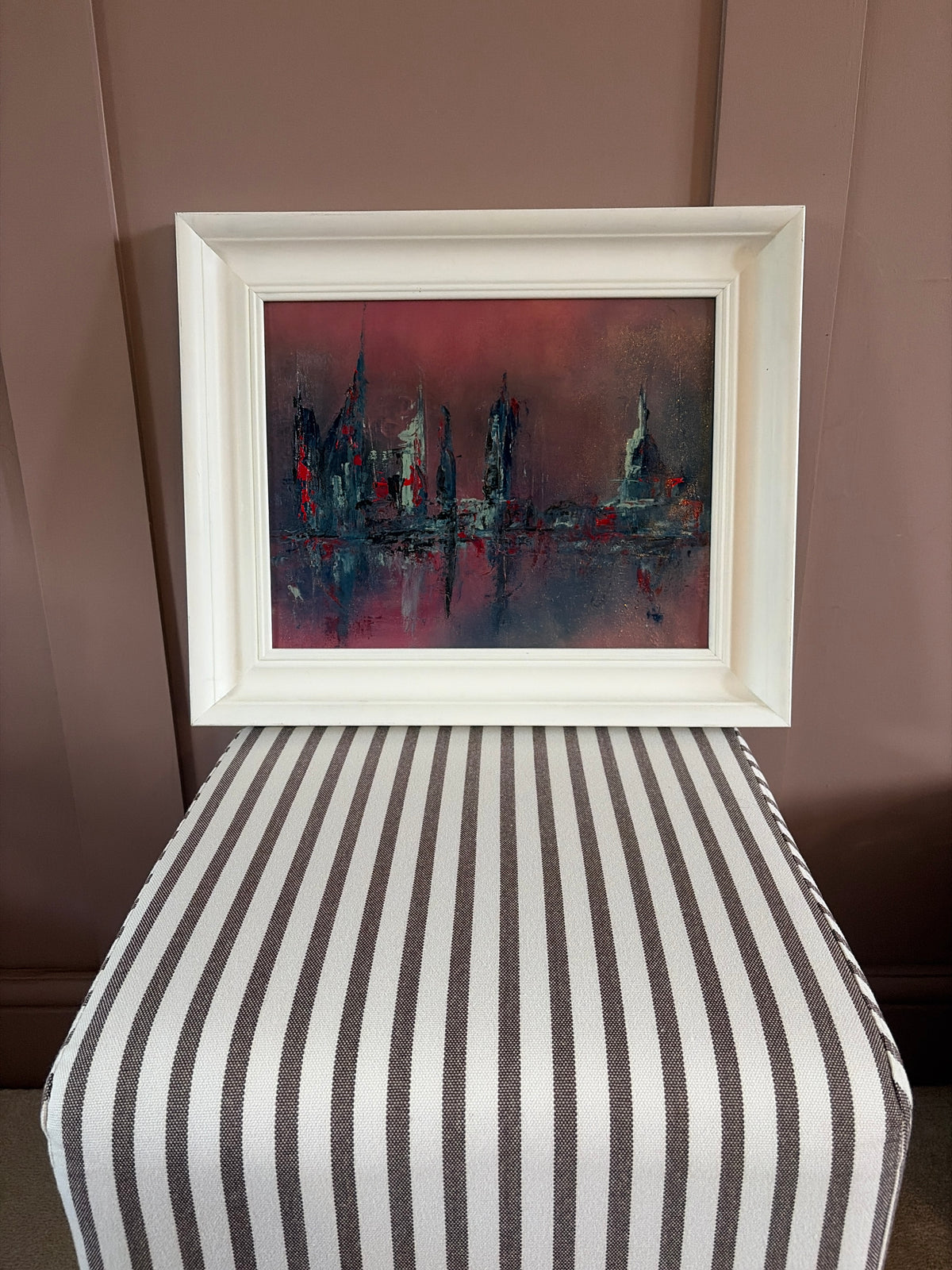 A framed abstract painting featuring a red and dark blue cityscape with reflections, displayed on a striped ottoman against a brown paneled wall.